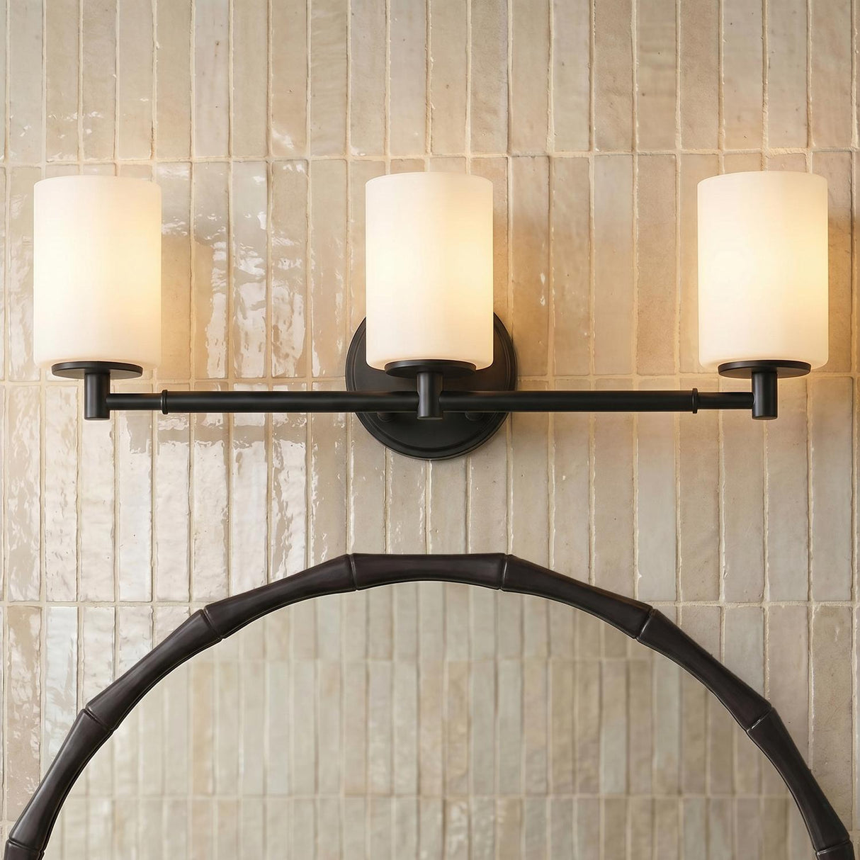 A contemporary bathroom features the Becca 3 Light Vanity, Matte Black with frosted glass shades mounted above a bamboo style mirror on a vertical beige tiled wall.