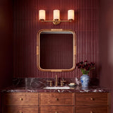 A contemporary bathroom with the Becca 3 Light Vanity, Aged Brass, featuring white frosted glass shades mounted above a rattan-trimmed mirror and burgundy fluted wall tiles.