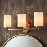 A modern bathroom featuring the Becca 3 Light Vanity, Aged Brass with frosted glass shades installed above a round gold-framed mirror on a dark textured wall.