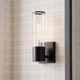 A modern farmhouse foyer featuring the Lea Wall Sconce, Matte Black with a clear glass cylinder and Edison bulb mounted on neutral plaid wallpaper beside a doorway.