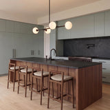 A modern kitchen with a matte black linear pendant featuring frosted glass globes over a wood island with black stone counters and sage green cabinetry.