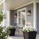 A craftsman porch with two matte black Paterson Outdoor Wall Light, Medium fixtures glowing beside a white window on gray siding, featuring white flowers in black planters.