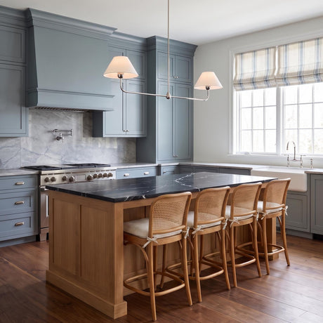 A transitional kitchen features the Holden 2 Shade Linear Pendant 57", Polished Nickel with white linen shades over a black marble island and blue cabinetry.