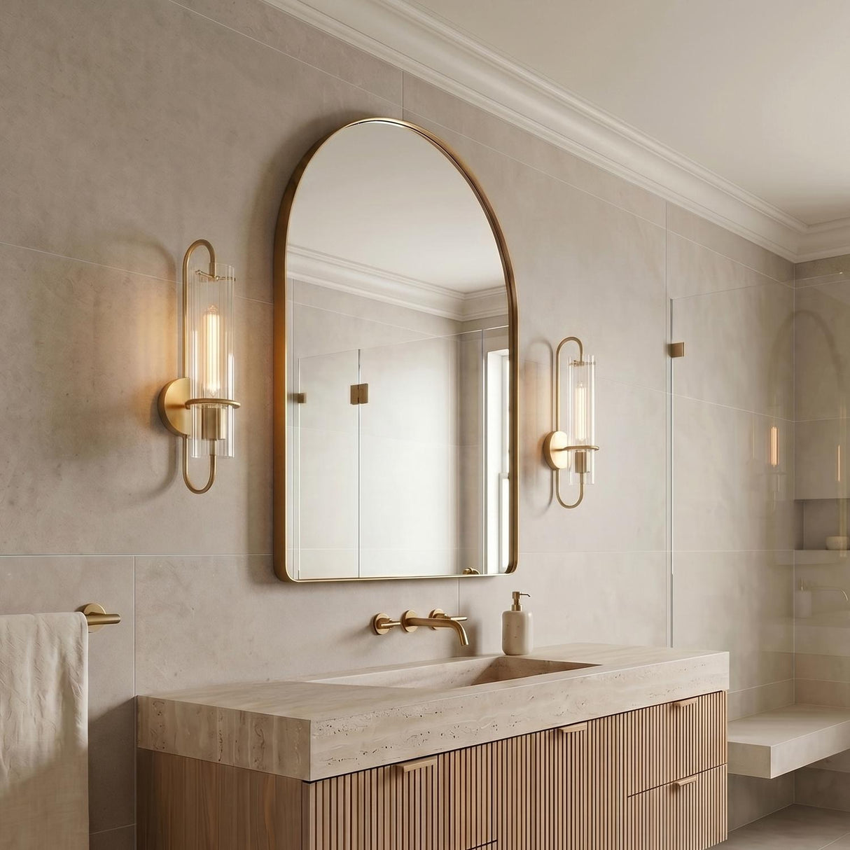 A transitional bathroom with two Camie Wall Sconce 20", Aged Brass fixtures flanking an arched mirror above a fluted wood vanity and light stone countertop.