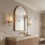 A transitional bathroom with two Camie Wall Sconce 20", Aged Brass fixtures flanking an arched mirror above a fluted wood vanity and light stone countertop.