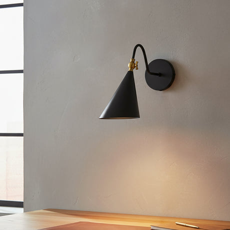 Faye Adjustable Metal Wall Sconce, Black with Aged Brass - A black cone-shaped wall sconce with aged brass accents is mounted on a textured gray wall above a wooden desk.