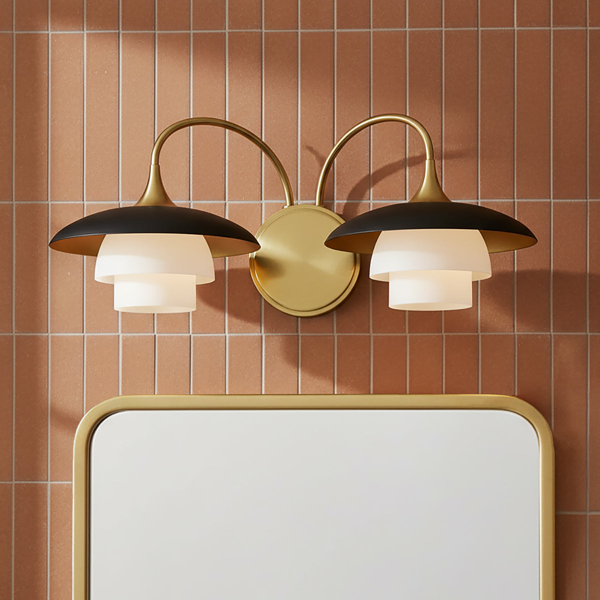 Wren 2 Light Wall Sconce, Matte Black and Aged Brass - A wall sconce with matte black shades and aged brass accents is mounted above a mirror with an aged brass frame, set against a backdrop of terracotta-colored tile.