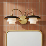 Wren 2 Light Wall Sconce, Matte Black and Aged Brass - A wall sconce with matte black shades and aged brass accents is mounted above a mirror with an aged brass frame, set against a backdrop of terracotta-colored tile.
