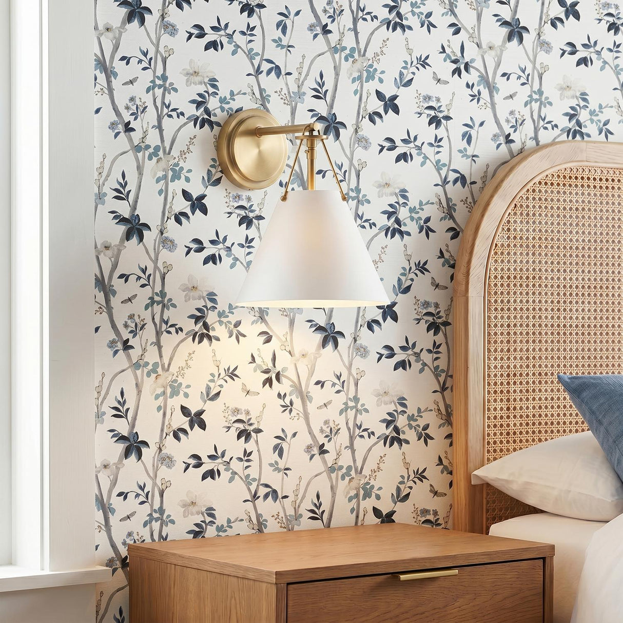 A transitional bedroom with the Bowie Wall Sconce, White and Brass, featuring a brushed brass finish mounted on floral wallpaper beside an arched rattan headboard.