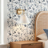 A transitional bedroom with the Bowie Wall Sconce, White and Brass, featuring a brushed brass finish mounted on floral wallpaper beside an arched rattan headboard.