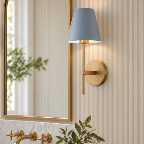 A transitional bathroom vanity features the Banks Wall Sconce, French Blue and Brass, mounted beside a brass arched mirror above a white marble countertop with striped wallpaper.