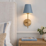 A coastal bedroom features the Banks Wall Sconce, French Blue and Brass mounted on a white wall beside a textured linen headboard and wooden nightstand.