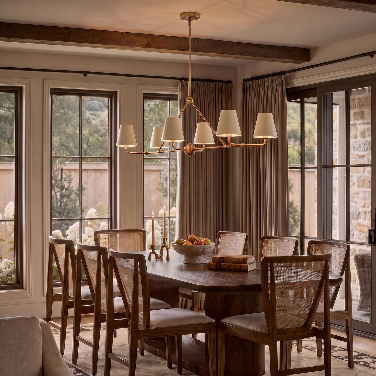 A modern farmhouse dining room with a brushed brass linear chandelier and white linen shades over a dark wood table with cane chairs and beamed ceilings.