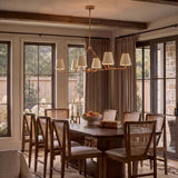 A modern farmhouse dining room with a brushed brass linear chandelier and white linen shades over a dark wood table with cane chairs and beamed ceilings.