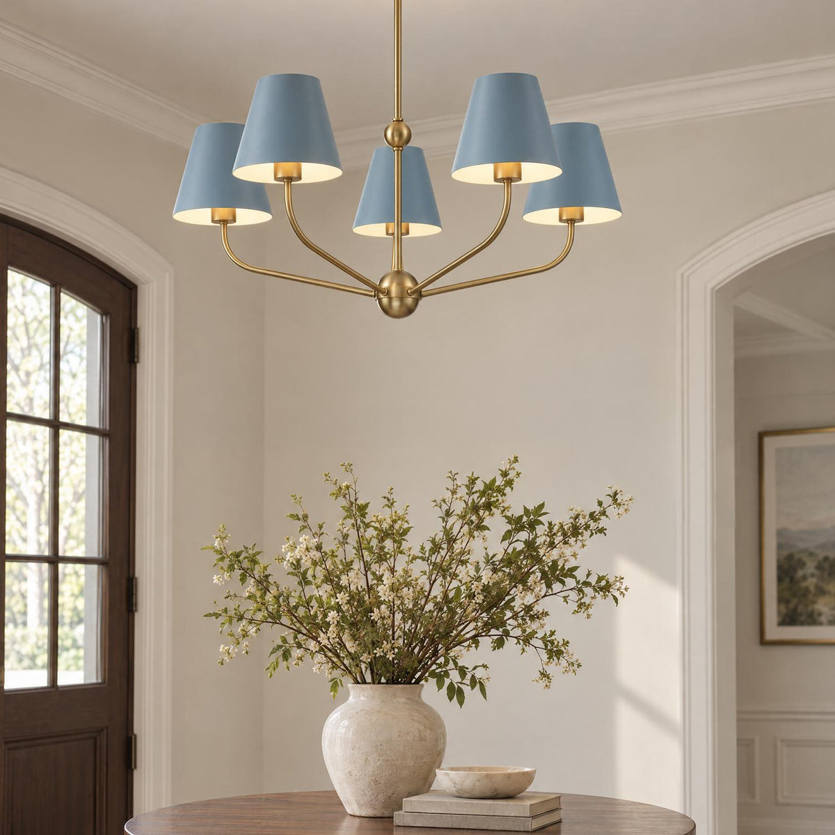 A transitional foyer features a brushed brass five-light chandelier with french blue shades suspended above a round wood entry table with a ceramic vase.
