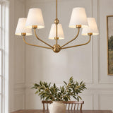 A transitional dining room featuring a brushed brass five light chandelier with white tapered fabric shades hanging above a wooden table against paneled white walls.