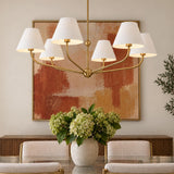 A transitional dining room features a brushed brass chandelier with white fabric shades suspended above a glass dining table with a green hydrangea centerpiece.