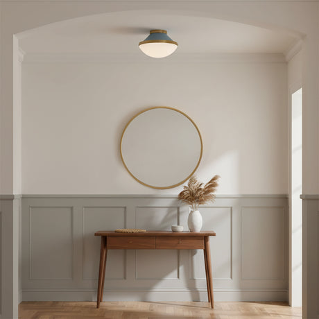 Blair Flush Mount 12, French Blue and Brass - A flush mount light fixture with a blue top and white shade is mounted on the ceiling above a gold-framed round mirror and a wooden console table with a vase of dried reeds.
