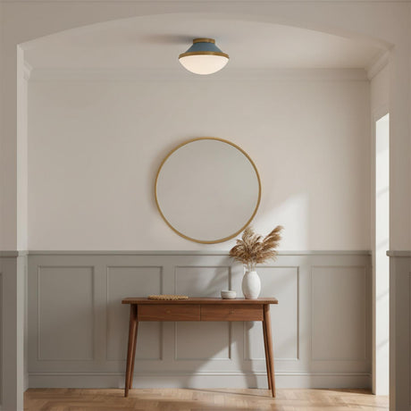 Blair Flush Mount 12, French Blue and Brass - A flush mount light fixture with a blue top and white shade is mounted on the ceiling above a gold-framed round mirror and a wooden console table with a vase of dried reeds.