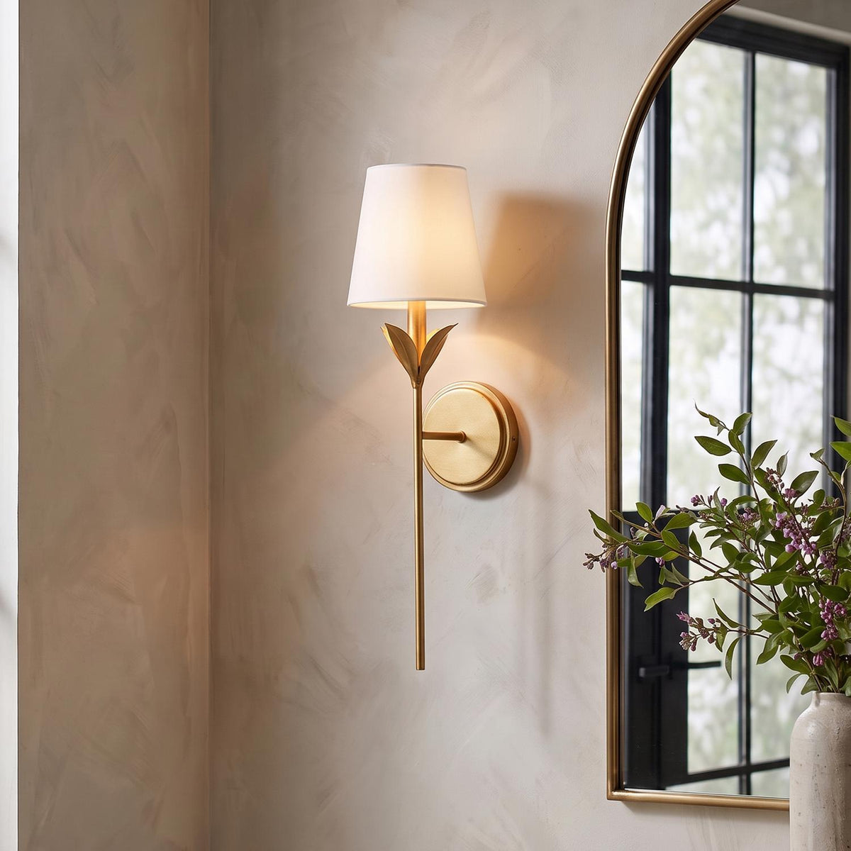 A transitional foyer feature wall showcasing an Alice Single Wall Sconce in antique gold with a white linen shade mounted beside an arched gold mirror.