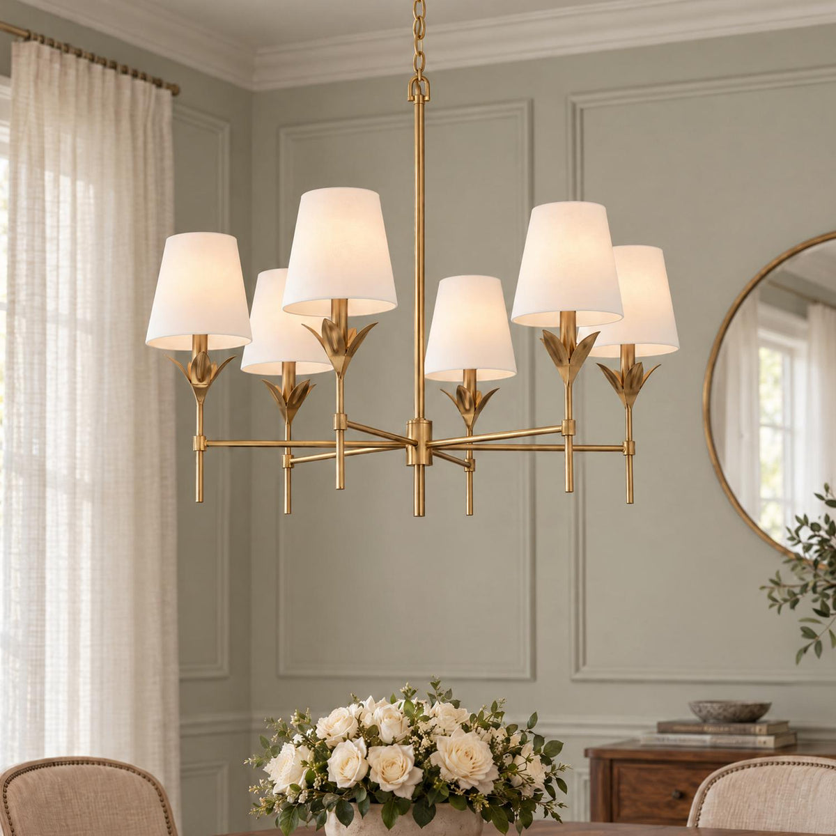 A transitional dining room featuring an antique gold six-light chandelier with white linen shades hanging above a wooden table with a white rose centerpiece.