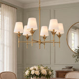 A transitional dining room featuring an antique gold six-light chandelier with white linen shades hanging above a wooden table with a white rose centerpiece.
