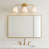 Hoyt 3 Light Vanity Light, Antique Gold with Opal Glass, shown above a single sink and mirror in a neutral decorated bathroom.