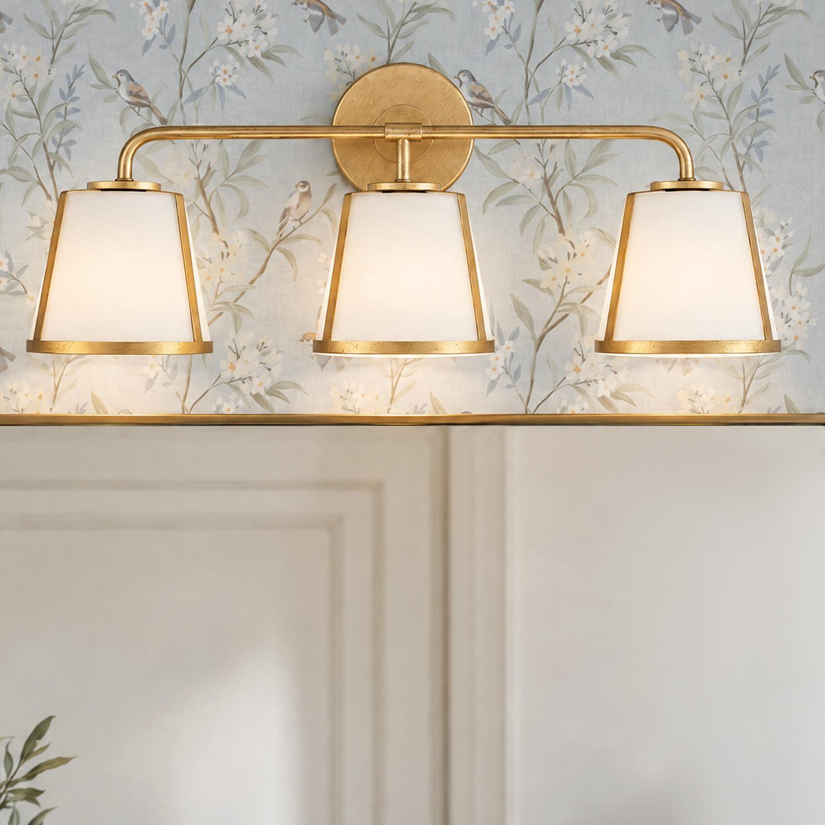 A transitional bathroom features the Hoyt 3 Light Vanity, Antique Gold with Opal Glass mounted above a wall mirror against floral botanical wallpaper.