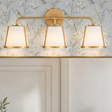 A transitional bathroom features the Hoyt 3 Light Vanity, Antique Gold with Opal Glass mounted above a wall mirror against floral botanical wallpaper.