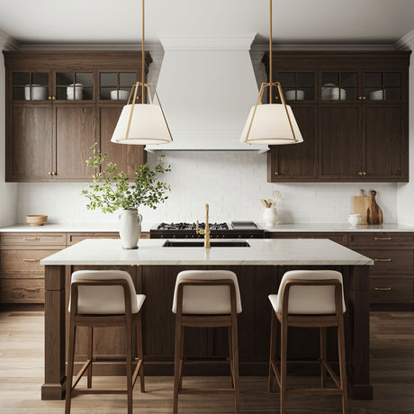 Two 20" wide Hoyt Pendant lights in Antique Gold with white shades are shown in a traditional kitchen, hung over a kitchen island, with dark walnut cabinetry and bright white stone countertops, with seating for three.