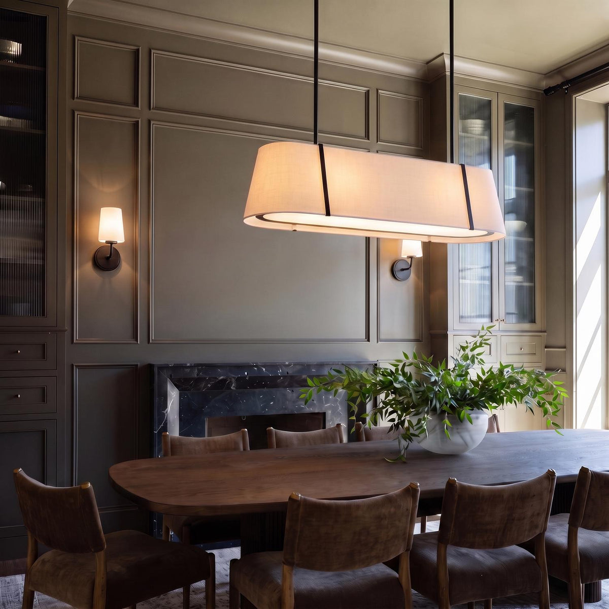 A transitional dining room featuring the Hoyt Linear Pendant 42", Black with a linen shade over a walnut table beside gray paneled walls and a marble fireplace. Two Wyatt Wall Sconces flank the fireplace on the wall.
