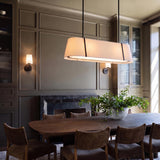A transitional dining room featuring the Hoyt Linear Pendant 42", Black with a linen shade over a walnut table beside gray paneled walls and a marble fireplace. Two Wyatt Wall Sconces flank the fireplace on the wall.