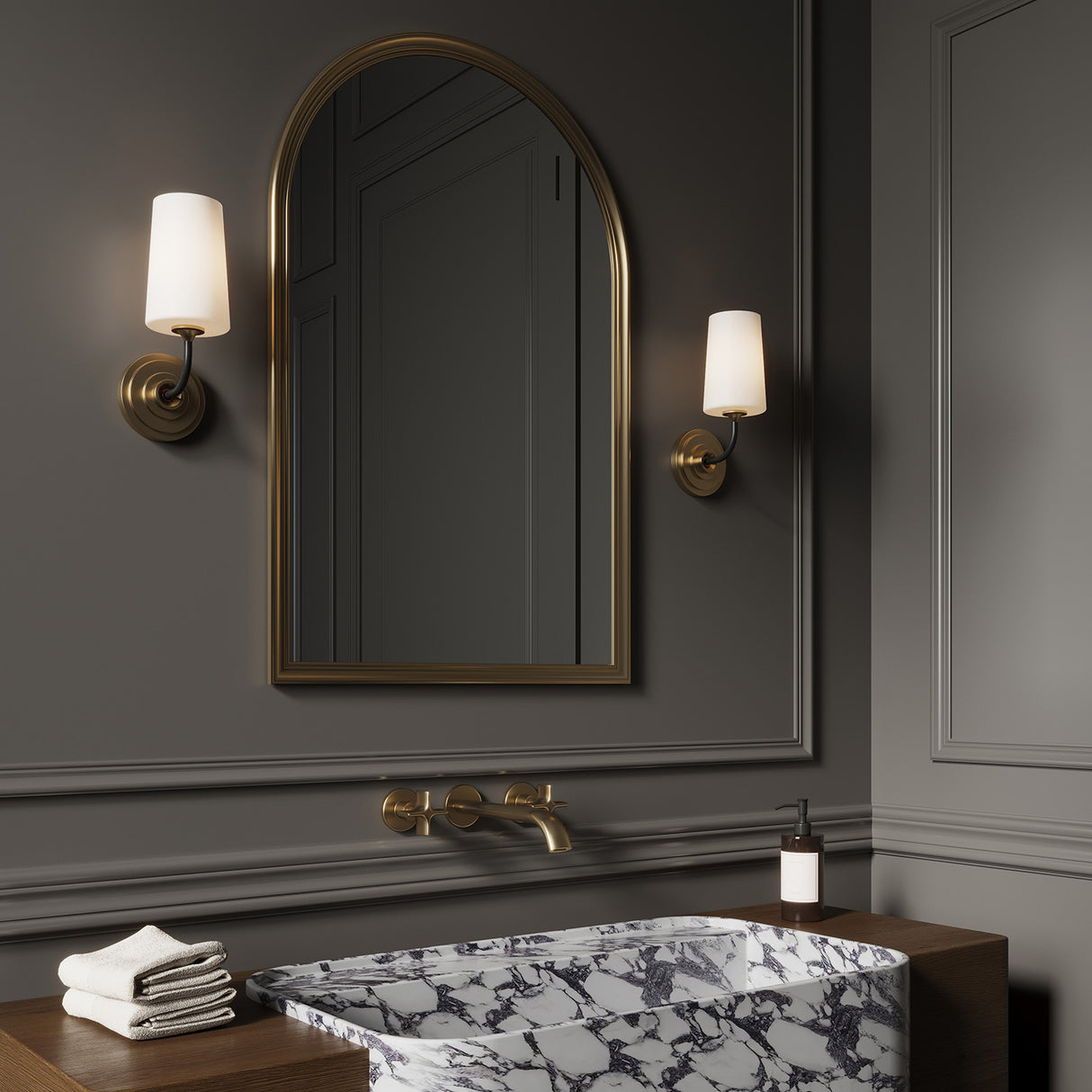 Two Logan wall sconces in black and brass with white opal glass shades, are flanking an arched brass mirror in a dark grey powder room, with classic styling and a marble slab sink.