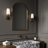 Two Logan wall sconces in black and brass with white opal glass shades, are flanking an arched brass mirror in a dark grey powder room, with classic styling and a marble slab sink.