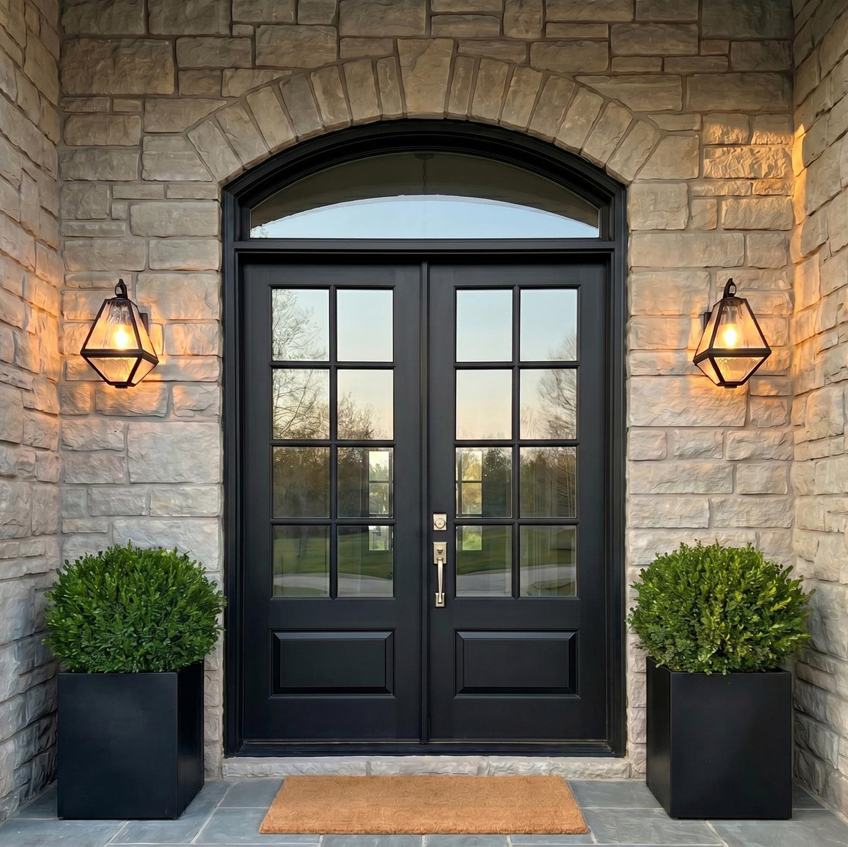 A traditional stone entry with matte black Emmett Outdoor Wall Lights flanking a black arched front door at twilight, accompanied by boxwood shrubs in planters.