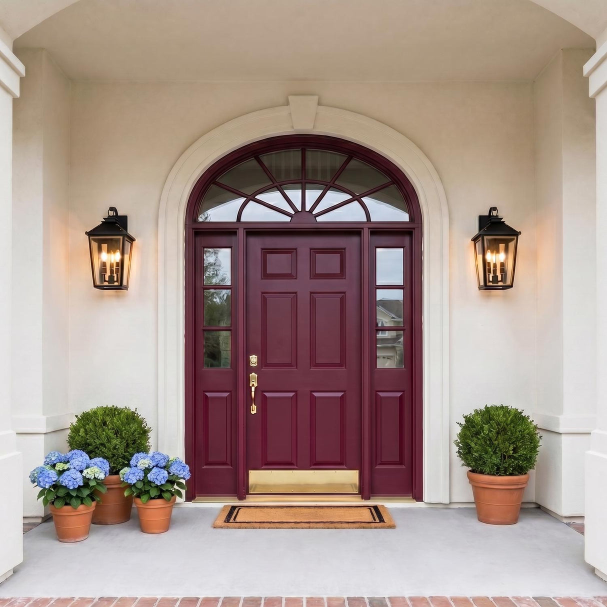 A Mediterranean home entry with two matte black Ellington Outdoor Wall Light fixtures flanking a burgundy front door on beige stucco with blue hydrangeas and boxwood.