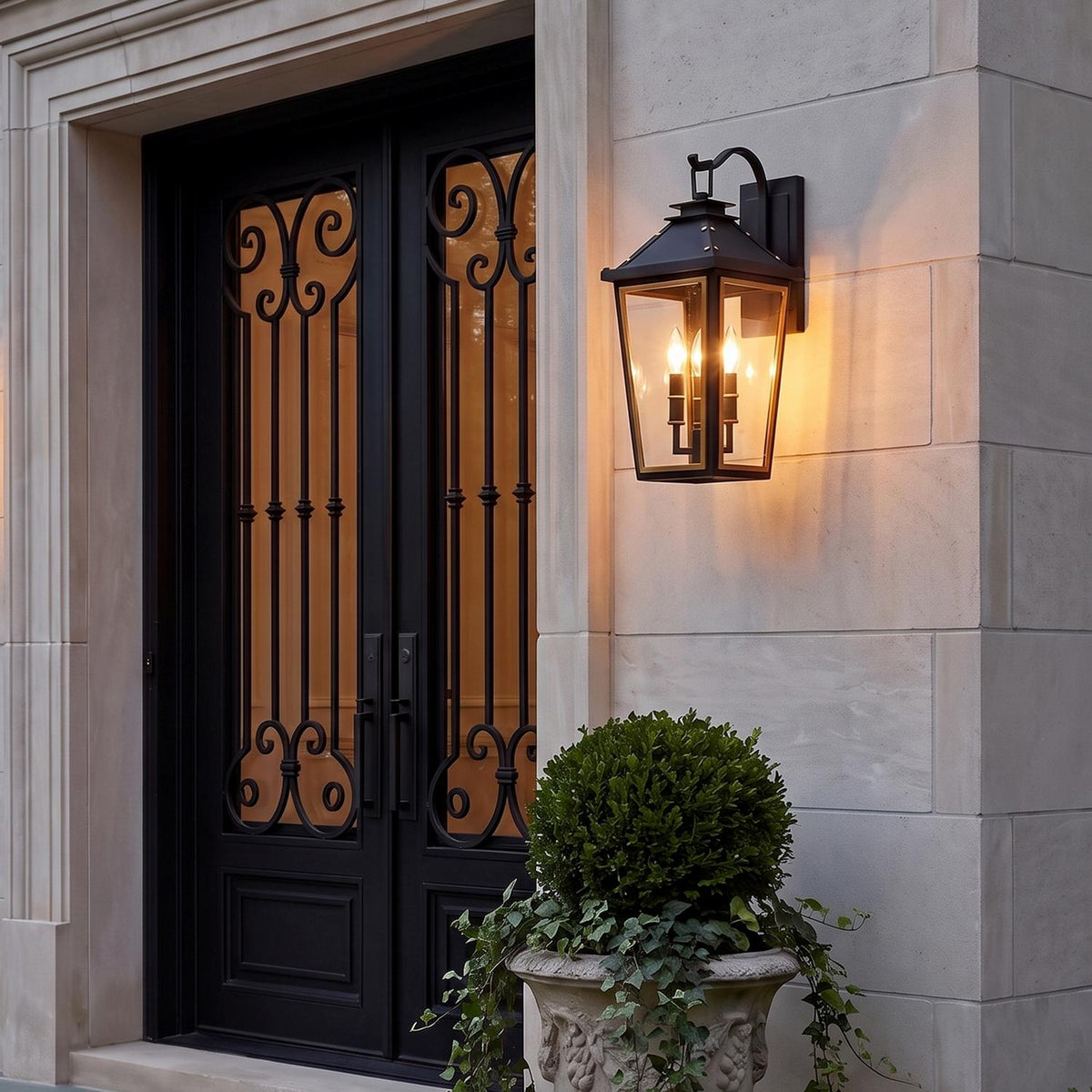 A traditional stone facade entry with an Ellington Outdoor Wall Light, Medium 19" Black and Textured Gold lantern glowing beside black wrought iron doors and potted boxwood.