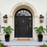 A Mediterranean stucco entryway featuring two matte black and textured gold wall lanterns flanking a dark arched front door with large potted elephant ear plants.