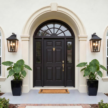 A Mediterranean stucco entryway featuring two matte black and textured gold wall lanterns flanking a dark arched front door with large potted elephant ear plants.
