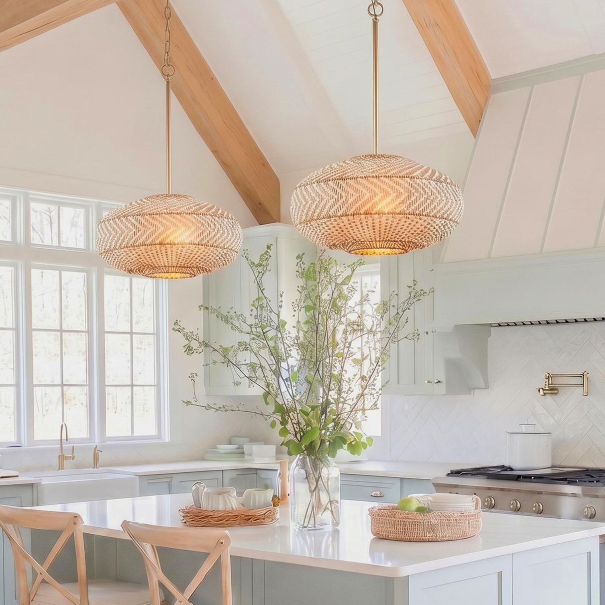 A coastal kitchen with two rattan and soft brass saucer pendants over a white island featuring light blue cabinets and wood beams on a vaulted ceiling.