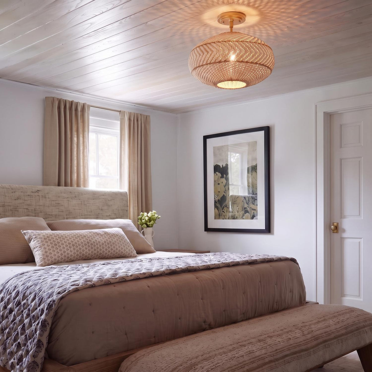 A coastal bedroom with a rattan and soft brass flush mount on a white wood-paneled ceiling above a bed with neutral linens and tan curtains.