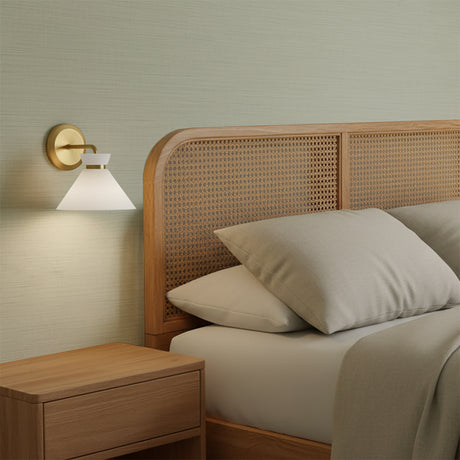 Cori Wall Sconce, Brushed Brass and Milk Glass - A brass and milk glass wall sconce next to a wooden bed frame.