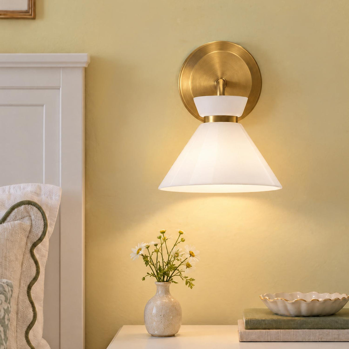 A transitional bedroom features the Cori Wall Sconce, Brushed Brass and Milk Glass mounted above a nightstand displaying a ceramic vase of daisies.