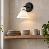 A modern kitchen featuring the Cori Wall Sconce, Matte Black and Milk Glass mounted on a glazed beige tile wall above a wooden shelf with ceramic dishes.