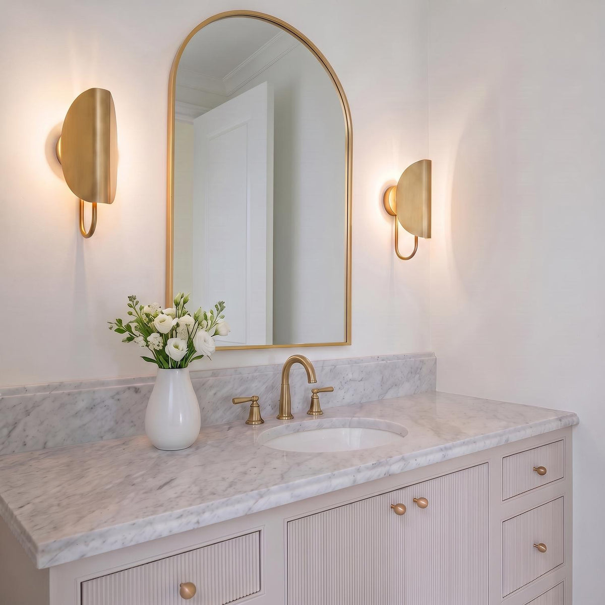 A contemporary bathroom with two Nico Wall Sconce, Antique Brass fixtures flanking an arched gold mirror above a white marble countertop and fluted taupe vanity.