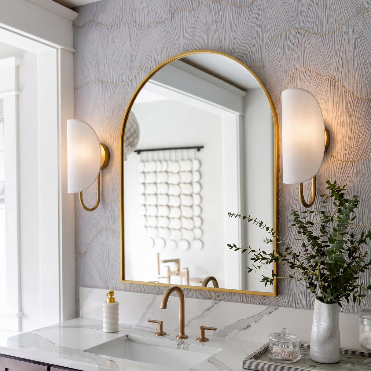 A transitional bathroom features two Nico Wall Sconces, Antique Brass with Fabric Shades, flanking an arched gold mirror above a white marble countertop and textured wallpaper.