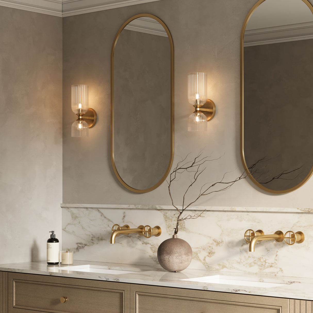 Elle Wall Sconce shown in brass with clear glass in a bathroom, flanking tall oval shaped vanity mirrors. 