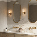 Elle Wall Sconce shown in brass with clear glass in a bathroom, flanking tall oval shaped vanity mirrors. 