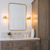 A transitional bathroom with two Elle Wall Sconce, Brushed Brass and Clear Glass fixtures flanking a gold mirror above a terrazzo countertop and reeded wood vanity.