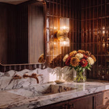 A mid-century bathroom featuring an Elle Wall Sconce, Brushed Brass and Clear Glass mounted on brown vertical tiles above a marble vanity and integrated sink.
